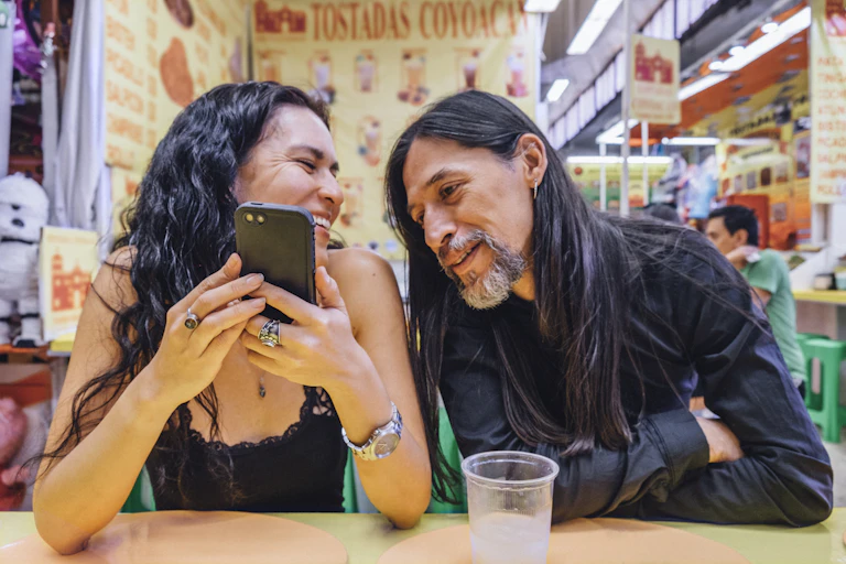A woman and a man seated side by side at a counter, the woman holds a phone and smiles at the man as he looks at the phone's screen