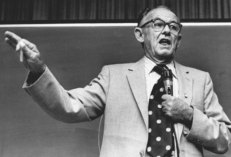 A black and white photograph shows a balding man giving a talk while wearing glasses, a suit and a polka-dot tie.