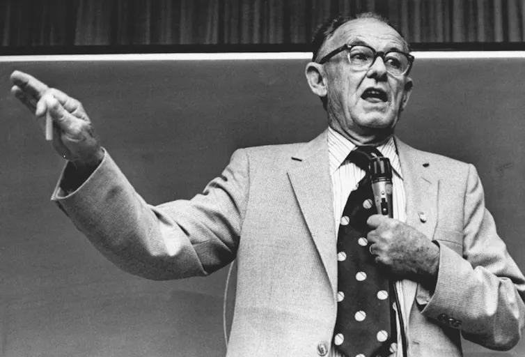 A black and white photograph shows a balding man giving a talk while wearing glasses, a suit and a polka-dot tie.