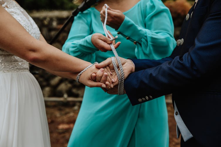A woman in a teal dress uses a silver cord to bind the hands of a person in a white dress and a person in a blue suit, who face each other.