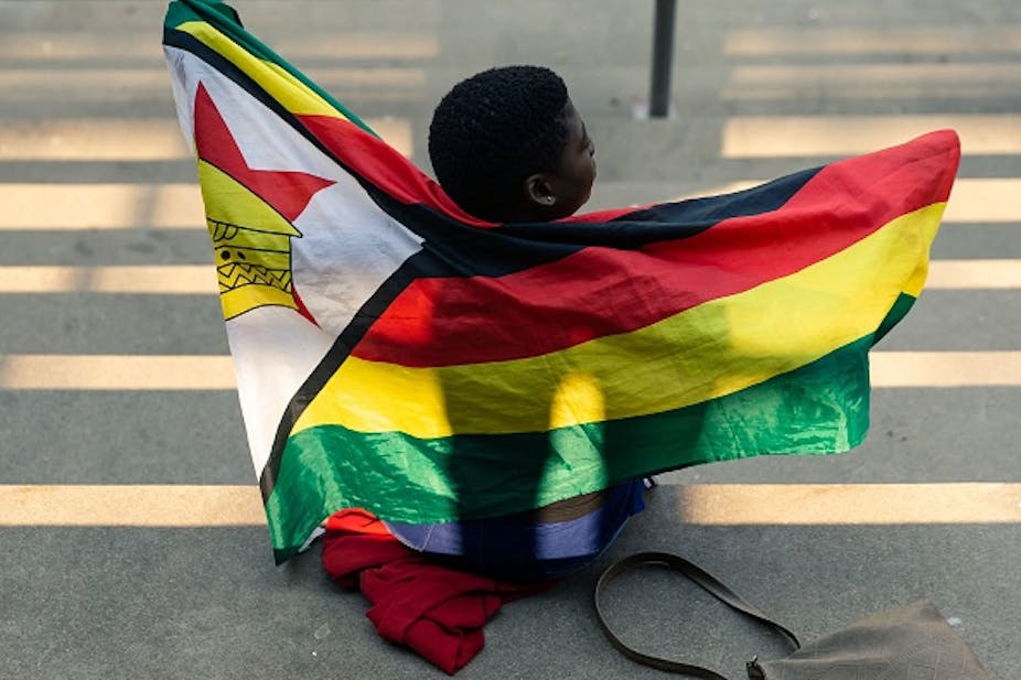 A child draped in Zimbabwe's national flag sits on the ground with her back to the camera. 