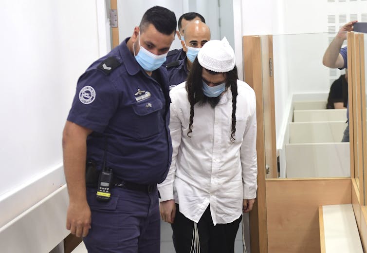 A man in a white shirt and cap and long braids bows his head as he walks in a courthouse with a police office beside him. Both wear masks