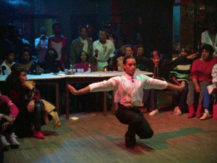 A man in a shirt squats to dance in front of a board of eager judges.