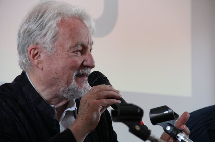 A man with gray hair and a white beard speaking into a microphone with a smile on his face