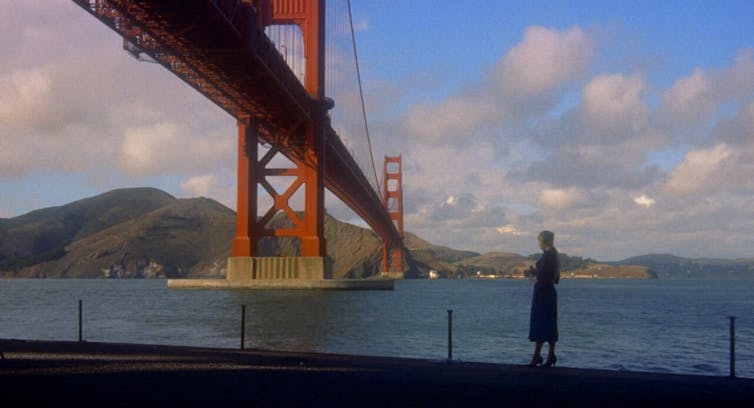 A woman walks along a river with a large red bridge, the Golden Gate Bridge, in the background