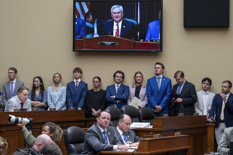 A group of young people in business dress, standing under a large screen with a gray haired man shown on it.