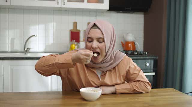 A woman wearing a hijab spoons oatmeal into her mouth.