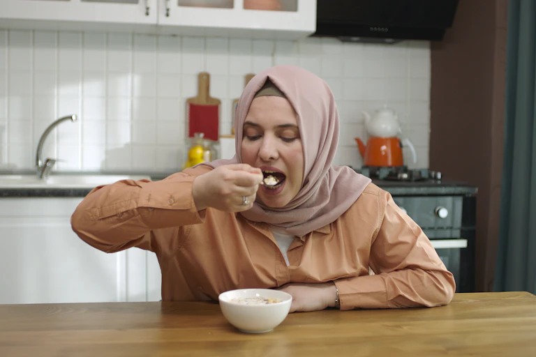 A woman wearing a hijab spoons oatmeal into her mouth.