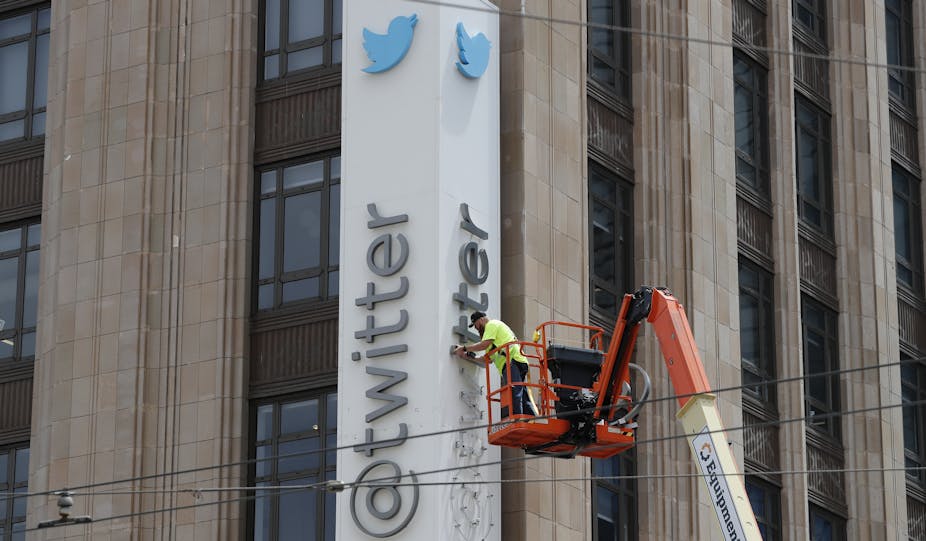 Building with long vertical sign showing blue bird logo and "@Twitter", a man on an orange crane is removing the letters.