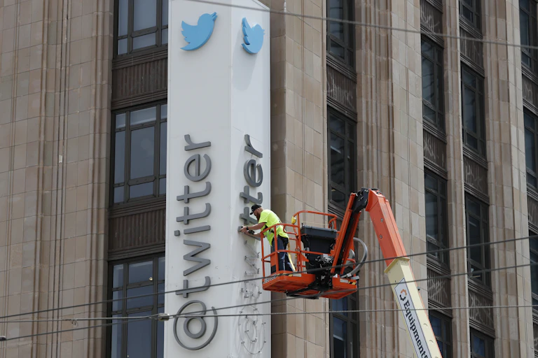 Building with long vertical sign showing blue bird logo and "@Twitter", a man on an orange crane is removing the letters.