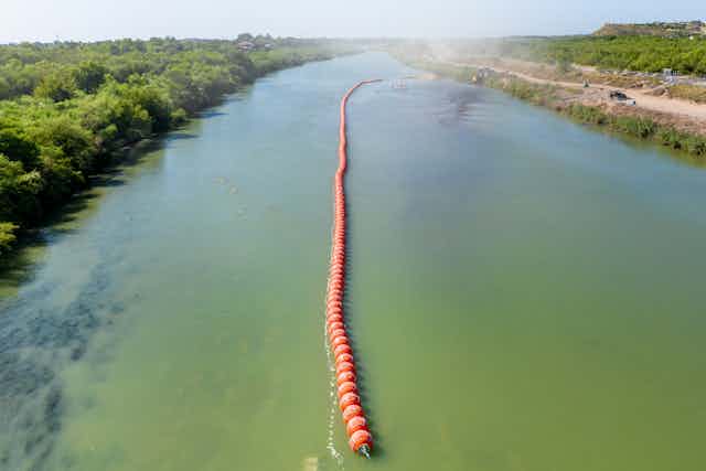 An aerial shot shows a long orange row of buoys in the middle of a green river. 