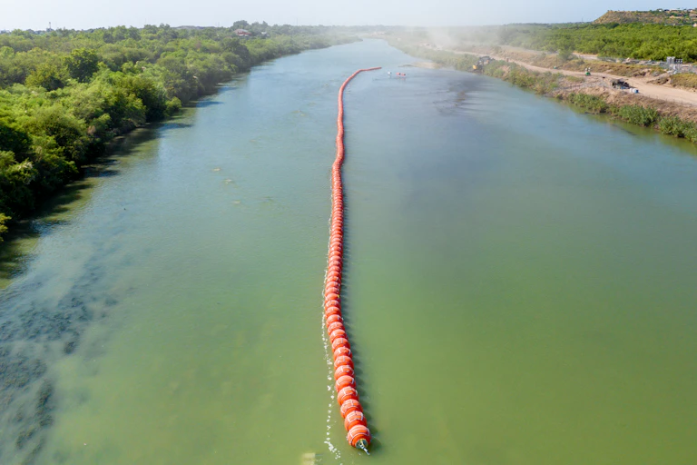 An aerial shot shows a long orange row of buoys in the middle of a green river.
