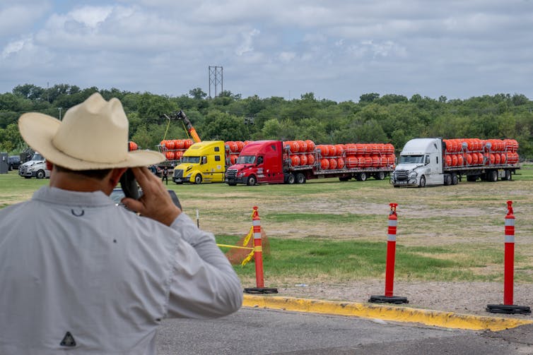 A person with a cowboy hat is seen from behind, talking on the phone as he looks at large trucks with big orange buoys lined up behind them.