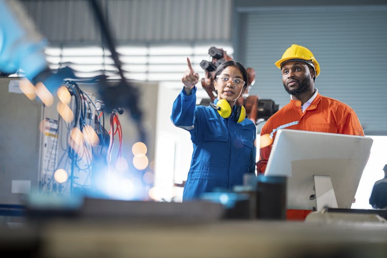 Two engineers standing in a factory, the one on the left, a woman, is pointing, while the one on the right, a man, wears a hard hat.