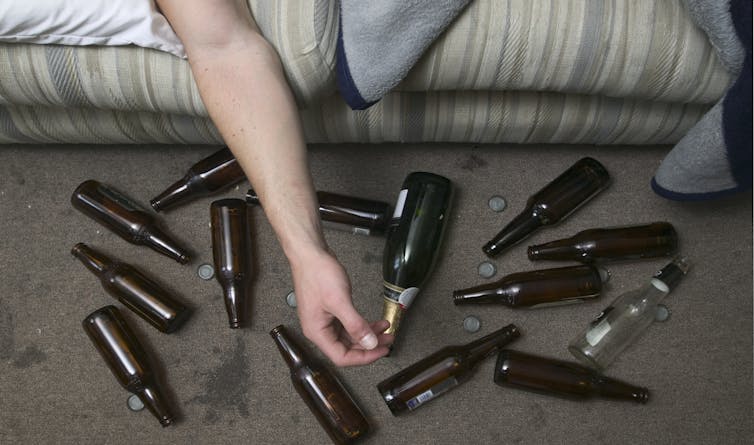 A bunch of beer bottles on the floor next to a person's arm hanging off couch.