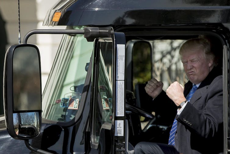A blond rotund man grimaces with his hands in fists in the cab of an 18-wheeler truck.