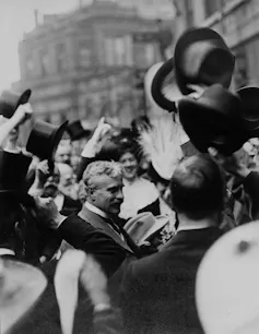 A man with grey hair and a moustache smiles among a crowd in a black-and-white photo.