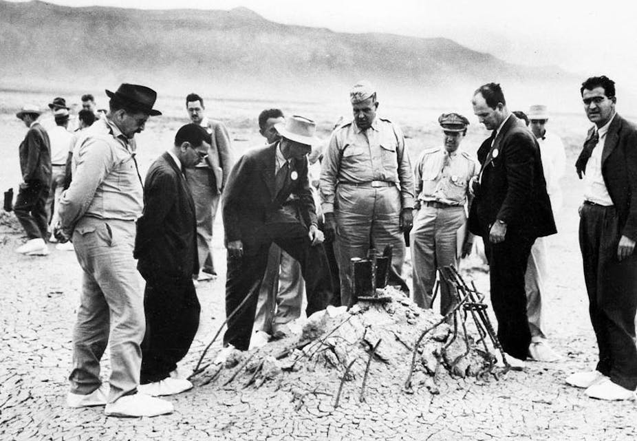 A black and white photograph of a group of men wearing suits or military uniforms, standing in the desert next to a pile of twisted metal remains from the first atomic explosion.