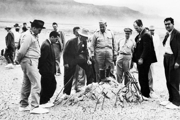A black and white photograph of a group of men wearing suits or military uniforms, standing in the desert next to a pile of twisted metal remains from the first atomic explosion.