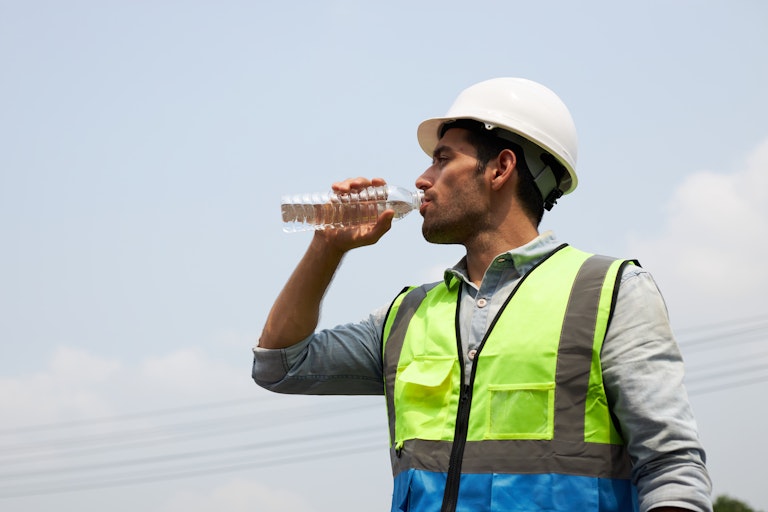 A man in a hardhat and hi-vis vest standing in the sun drinking a bottle of water