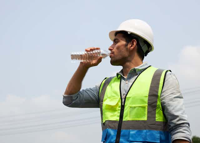 A man in a hardhat and hi-vis vest standing in the sun drinking a bottle of water