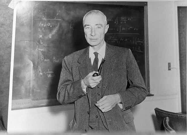 A gaunt man in a three-piece suit, holding a pipe, stands in front of a blackboard.