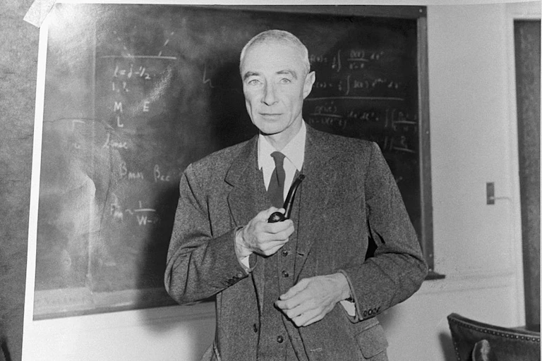 A gaunt man in a three-piece suit, holding a pipe, stands in front of a blackboard.