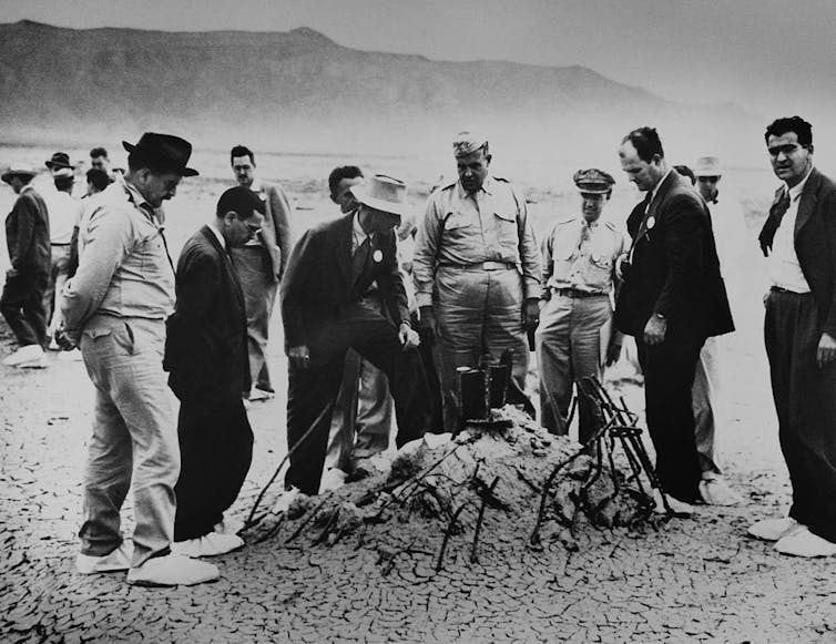 Eine Handvoll Männer in Anzügen und Militäruniformen stehen in der Wüste und betrachten einen Haufen verbrannten Metalls