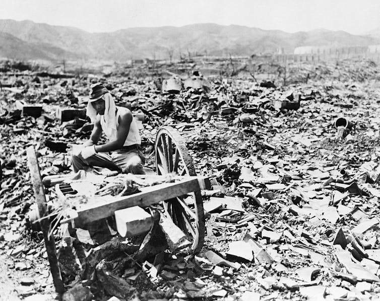A man with white fabric over his head sits on a cart amid a desolate, ruined city landscape.