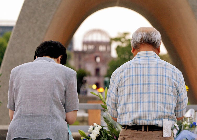 A woman and man with their backs to the camera bow as if in prayer before a concrete structure.