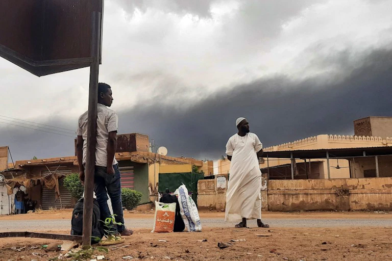 Two men stand by a roadside with bags by their feet as a plume of black smoke billows behind them