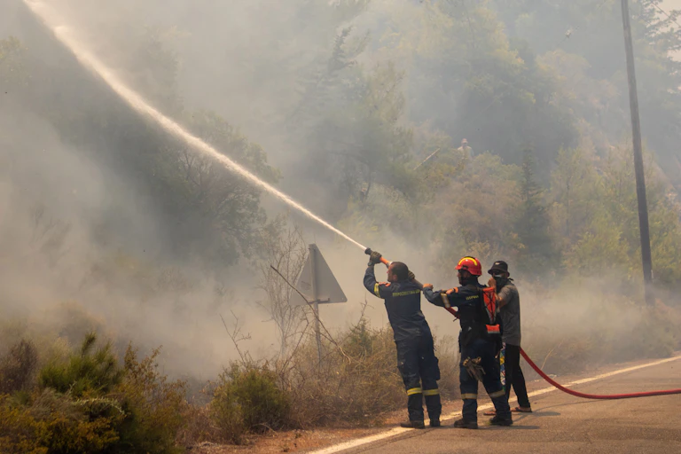 Three firefighters direct a jet of water into burning roadside vegetation.