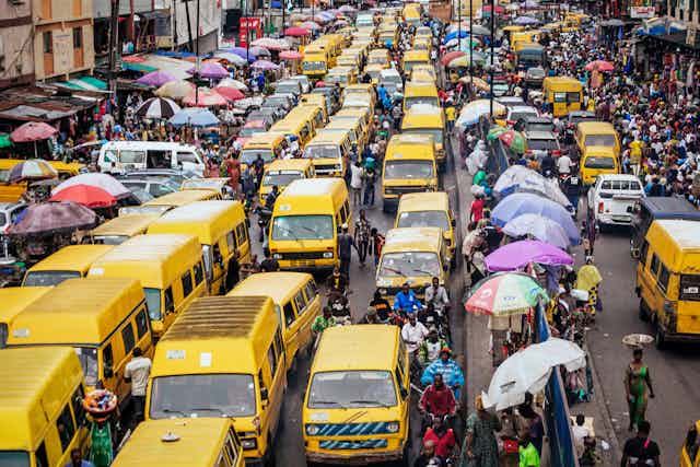 A busy street in Lagos, jam-packed with yellow minibus vehicles, commuters on the outskirts and a row of umbrellas with people selling goods in the middle of the streets.