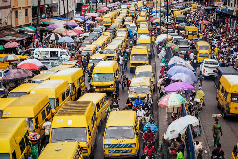 A busy street in Lagos, jam-packed with yellow minibus vehicles, commuters on the outskirts and a row of umbrellas with people selling goods in the middle of the streets.