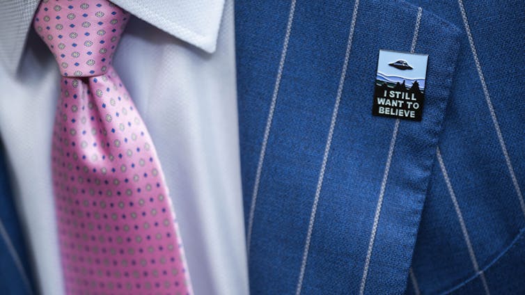 A close-up shot of a blue striped suit and pink tie with a rectangular pin that has a UFO on it and the words 'I still want to believe'