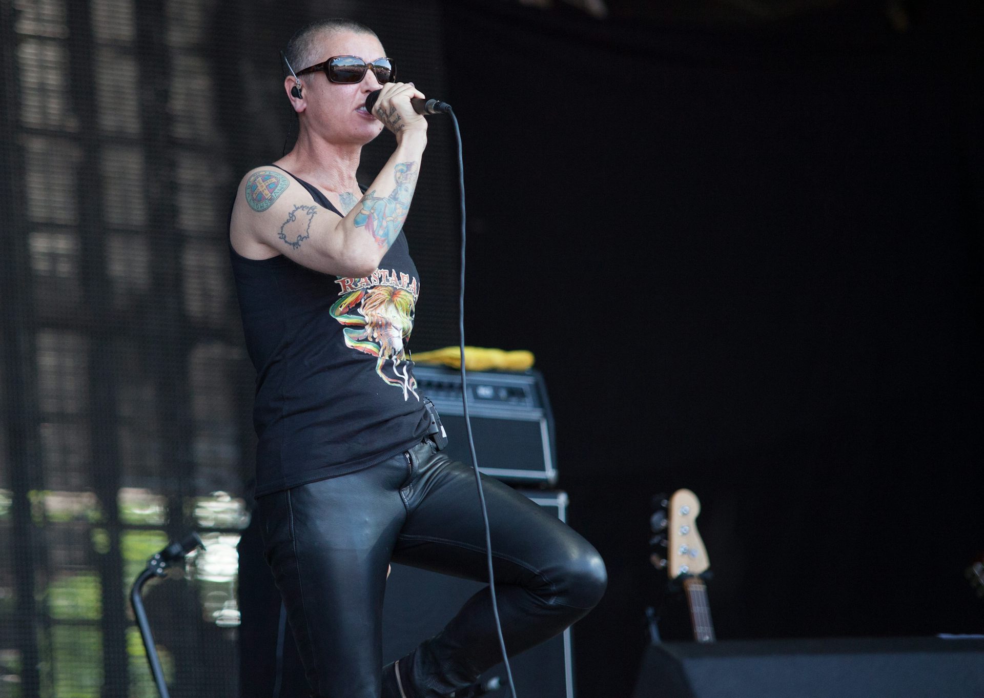 A tattooed skinhead woman singing on stage wearing black leather and sunglasses.