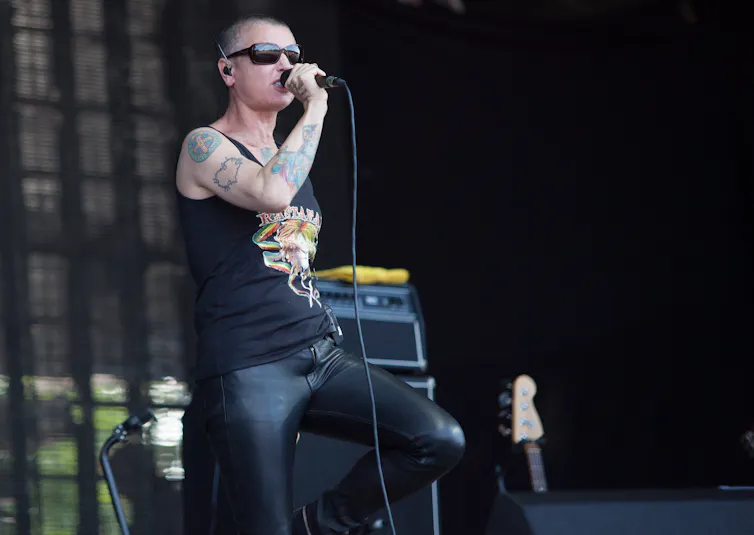 A tattooed skinhead woman singing on stage wearing black leather and sunglasses.
