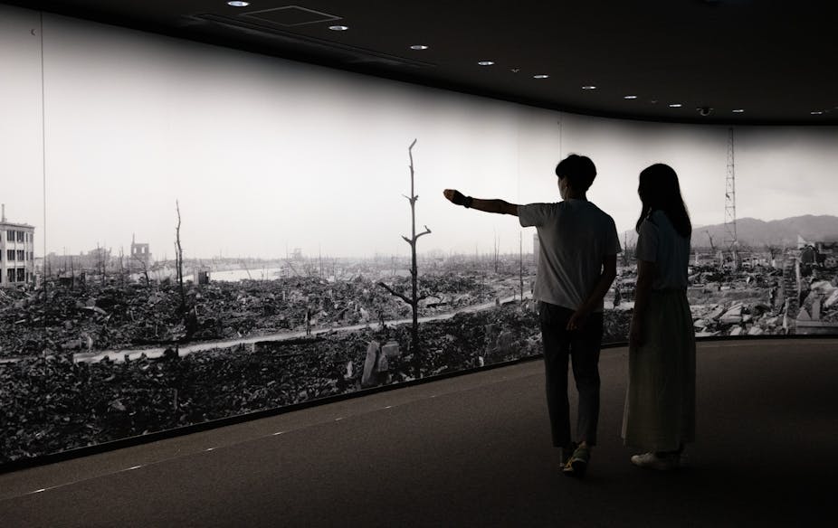Two people are seen with their backs to the camera in front of a long, floor to ceiling sized photo that shows a destroyed area and bare trees.