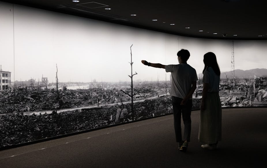 Two people are seen with their backs to the camera in front of a long,  floor to ceiling sized photo that shows a destroyed area and bare trees.