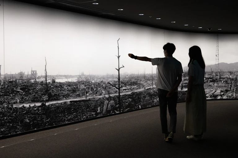 Two people are seen with their backs to the camera in front of a long, floor to ceiling sized photo that shows a destroyed area and bare trees.