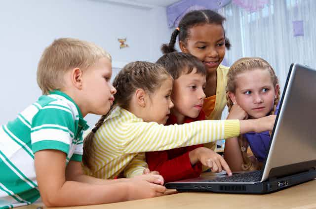 Varios niños observan la pantalla de un ordenador mientras una niña señala algo en ella.