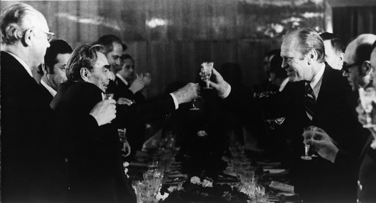 A black and white photo shows two men toasting each other, surrounded by other men at a table.