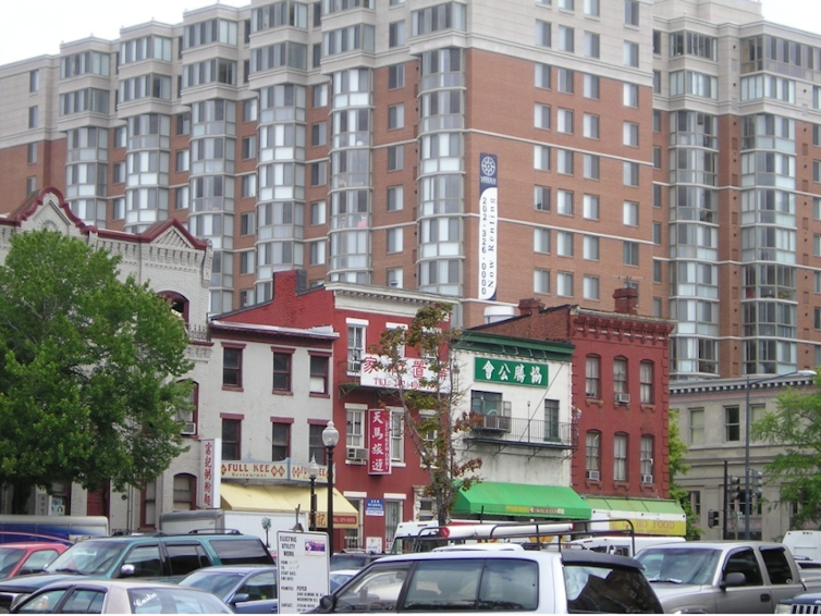 View of Chinese storefronts, with a large apartment building in the background and cars in the foreground