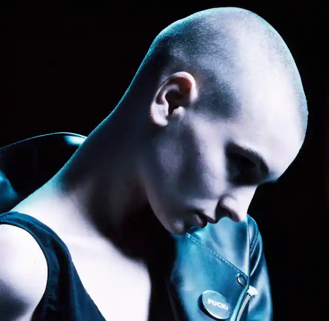A young women with a skinhead wearing a leather jacket off the shoulders spotlit against a black background.