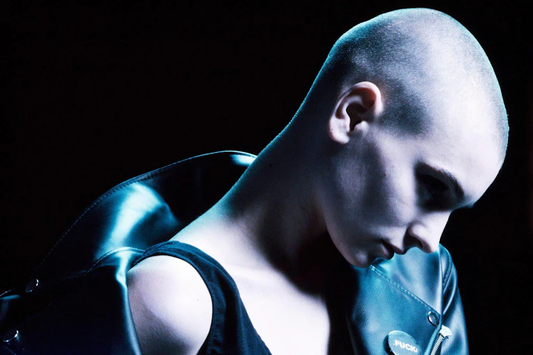 A young women with a skinhead wearing a leather jacket off the shoulders spotlit against a black background.