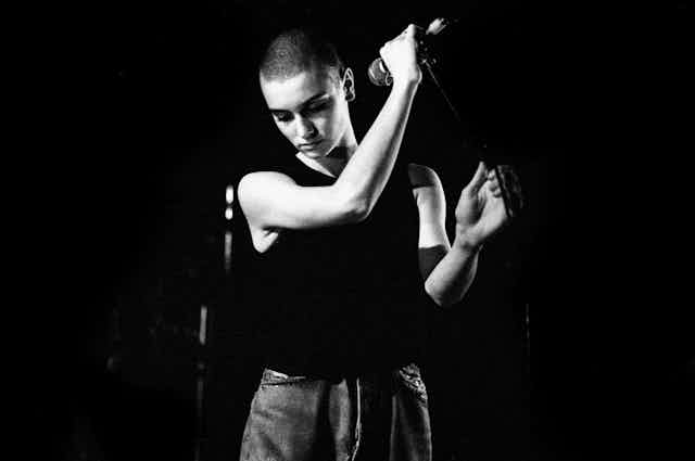A black and white photo centers on a woman with a shaved head, looking down in thought, who holds a microphone in an upraised hand.