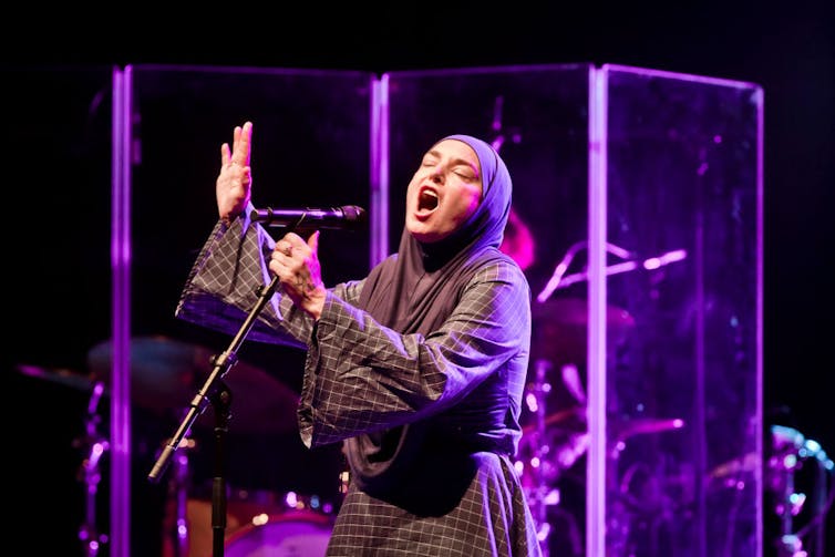 A woman in a checked robe and headdress sings passionately in front of purple lights.