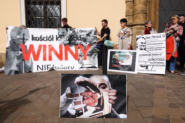 Une poignée de personnes tiennent des pancartes de protestation rouges, blanches et noires devant un bâtiment, avec une grande photo d’une femme déchirant une photographie devant eux