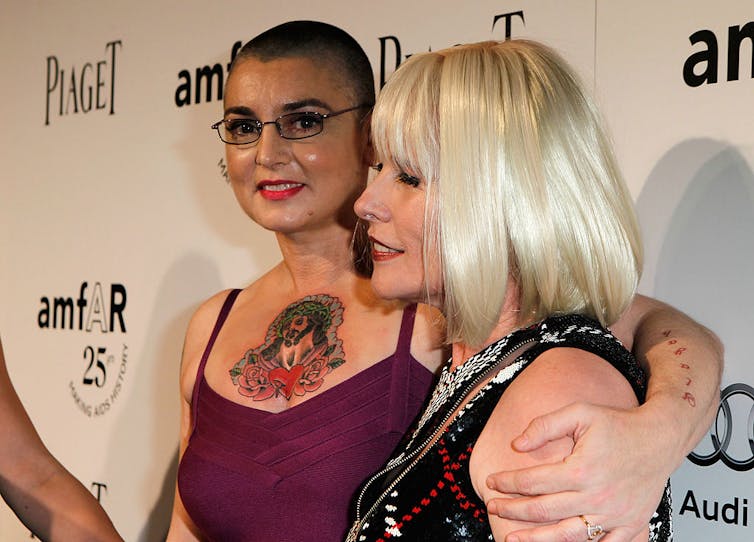 Une femme en robe violette, au crâne rasé et au grand tatouage coloré, embrasse une femme blonde