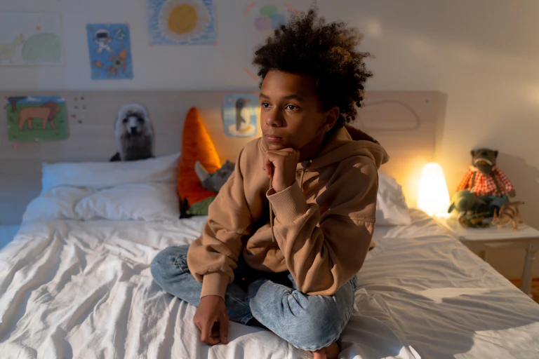 A boy sits on his bed, with a pensive expression.